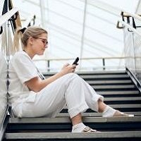 Foto av en kvinna som sitter i en trapp i vårdkläder med en telefon som hon tittar på.